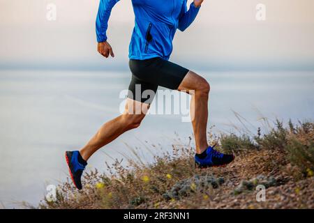 il corridore maschio ravvicinato corre in salita su trail con giacca blu e tights neri, sfondo di cielo e mare Foto Stock