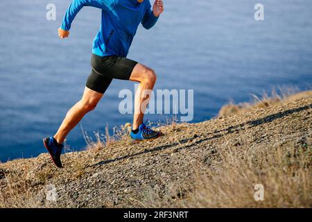l'atleta runner inizia a correre in montagna in salita con giacca blu e tights neri, sfondo di mare Foto Stock
