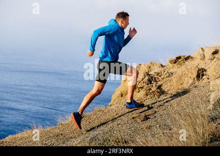 atleta runner che corre in montagna in salita con giacca blu e tights neri, sfondo di mare Foto Stock
