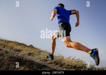 atleta runner che corre in montagna in salita con camicia blu e tights neri, sfondo di cielo Foto Stock
