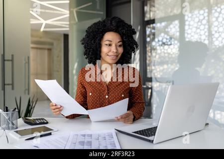 Giovane finanziatore di successo lavora con documenti e contratti all'interno dell'ufficio, soddisfatto dei risultati delle attività finanziarie, esamina conti, rapporti e contratti da donna d'affari, laptop. Foto Stock