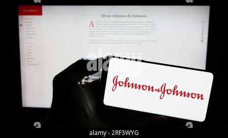 Persona in possesso di un cellulare con il logo della società statunitense Johnson and Johnson (JNJ) sullo schermo davanti alla pagina Web aziendale. Concentrarsi sul display del telefono. Foto Stock