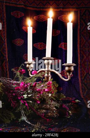 Un candelabro a tre ramificazioni con candele accese di fronte a un arazzo sbiadito, che illumina un bouquet di rose morenti e altri fiori Foto Stock