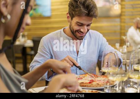 Giovane trentenne, che indossa una camicia, sorride mentre taglia la pizza con il coltello e la forchetta in una pizzeria del cortile. Un primo piano sfocato di una donna, un Foto Stock