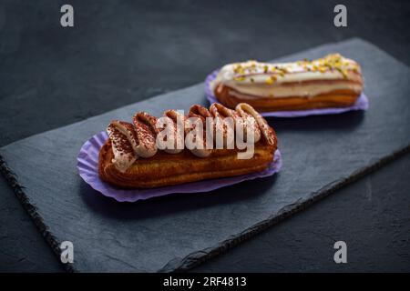 Due deliziose torte eclair - tiramisù e pistacchio aromatizzati ricoperti di panna, glassa e noci tritate, su sfondo nero Foto Stock