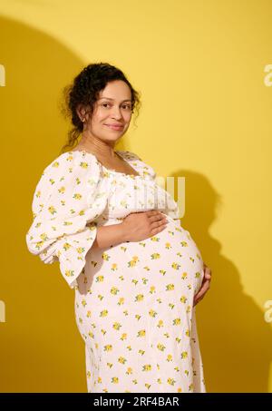 Ritratto in studio di una bella donna multi-etnica con i capelli ricci, vestita con un vestito bianco estivo, sorridente guardando la macchina fotografica, tenendola in mano Foto Stock