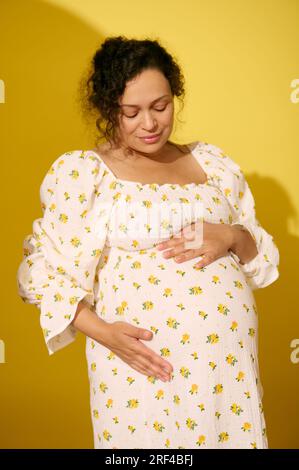 Bella donna incinta dai capelli ricci multietnici con elegante abito da sole estivo, accarezza delicatamente la pancia, sorride guardando la macchina fotografica, isolata sul giallo Foto Stock