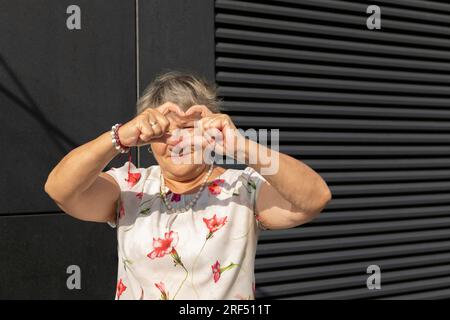 Carina anziana vecchia donna che fa Una forma di cuore con le sue mani e le sue dita. Bella donna reale 70 Yo matura con capelli grigi all'aperto, parete scura accesa Foto Stock