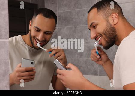 Uomo felice che usa lo smartphone mentre si lavano i denti in bagno. Dipendenza da Internet Foto Stock