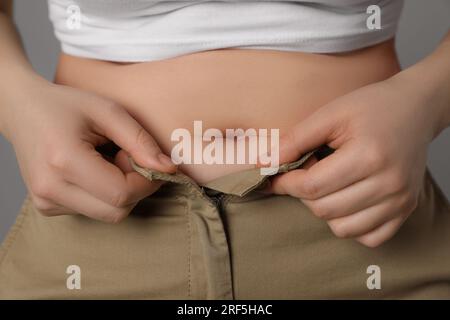 Donna in sovrappeso che cerca di indossare pantaloni stretti su sfondo grigio, primo piano Foto Stock