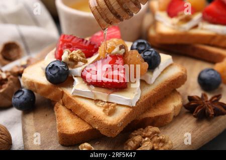 Versare il miele su gustosi panini con formaggio brie, frutti di bosco e noci sul tavolo, primo piano Foto Stock