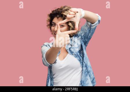 Ritratto di un'attraente fotografa concentrata con capelli ricci che indossa una camicia blu che guarda la fotocamera attraverso il fotogramma delle dita. Riprese in studio per interni isolate su sfondo rosa. Foto Stock