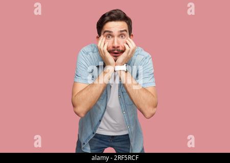 Ritratto di un uomo barbuto sbalordito in camicia blu casual in piedi con le mani sulle guance, guardando la fotocamera con grandi occhi. Riprese in studio per interni isolate su sfondo rosa. Foto Stock