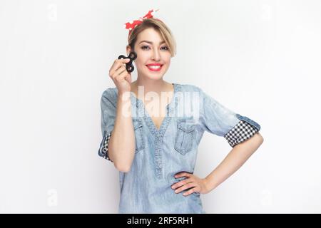 Ritratto di una donna bionda sorridente allegra e gioiosa che indossa una camicia in denim blu e un archetto rosso che regge lo spinner, tiene la mano sui fianchi. Riprese in studio in interni isolate su sfondo grigio. Foto Stock