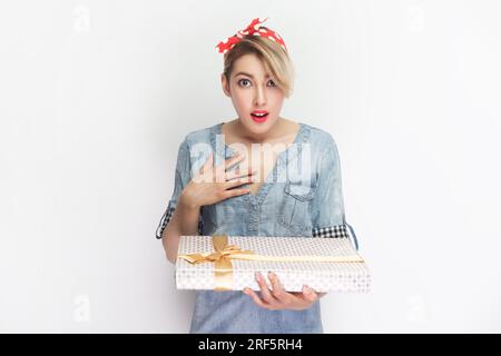 Il ritratto di una donna bionda sorpresa che indossa una camicia in denim blu e un cerchietto rosso che regge una scatola regalo, ottiene un regalo inaspettato. Riprese in studio in interni isolate su sfondo grigio. Foto Stock