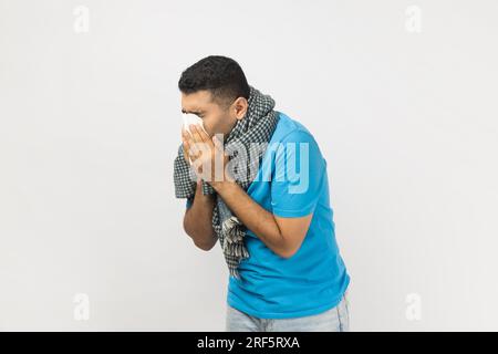 Ritratto di uomo malato e non rasato che indossa una maglietta blu e avvolto in una sciarpa calda starnuto, con naso che cola, ha grippa. Riprese in studio in interni isolate su sfondo grigio. Foto Stock