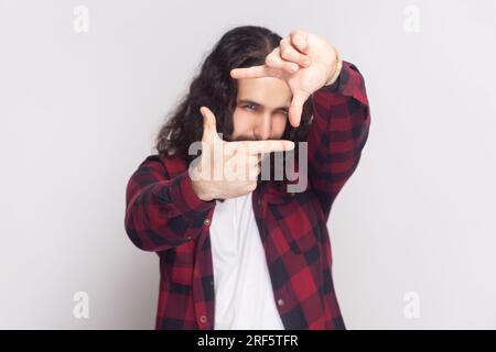 L'uomo barbuto con lunghi capelli ricci e camicia rossa a scacchi ha preso ispirazione immagina calda per catturare scatti interessanti che sorride volentieri il gesto dell'inquadratura. Riprese in studio in interni isolate su sfondo grigio. Foto Stock