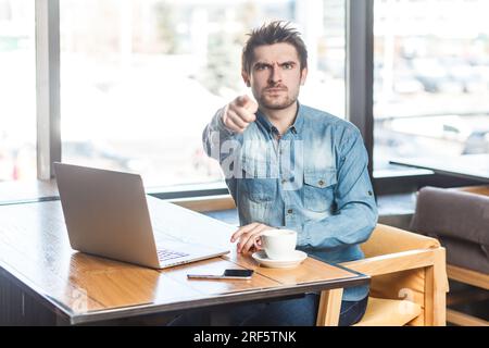 Ritratto di un giovane uomo tagliente e bello freelance con una camicia blu jeans che lavora su un computer portatile, che punta alla fotocamera e ti seleziona. Foto al coperto vicino a una grande finestra, sfondo del caffè. Foto Stock