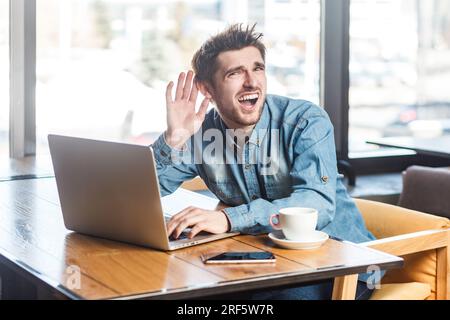 Ritratto di un uomo barbuto freelance in camicia blu jeans che lavora su un computer portatile, che ha problemi con l'altoparlante del microfono, tiene la mano vicino all'orecchio, cercando di sentire. Foto al coperto vicino a una grande finestra, sfondo del caffè. Foto Stock