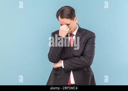 Ritratto di un uomo triste che piange con i baffi in piedi con la testa giù, che sente una brutta notizia, indossa un vestito nero con cravatta rossa. Riprese in studio in interni isolate su sfondo azzurro. Foto Stock