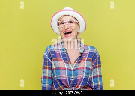Ritratto di una donna anziana che ride con camicia a scacchi, cappello e occhiali da vista molto felice, sorridente con ampio sorriso che mostra il suo umore perfetto. Riprese in studio in interni isolate su sfondo giallo. Foto Stock
