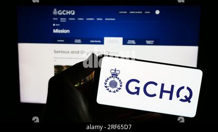 Persona che possiede il cellulare con il logo della sede centrale delle comunicazioni governative (GCHQ) sullo schermo davanti alla pagina Web. Concentrarsi sul display del telefono. Foto Stock