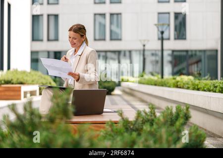 Analista donna che lavora con documenti e utilizza un notebook seduto all'esterno su un edificio moderno Foto Stock