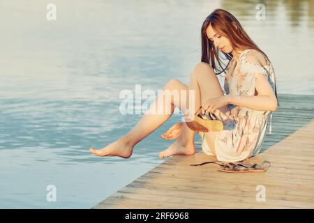 Una giovane donna con un abito da sole si siede sul bordo del molo e si toglie i sandali per immergersi in acqua Foto Stock