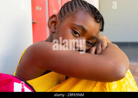 Ritratto di una triste studentessa afroamericana con una borsa della scuola seduta al muro nel corridoio della scuola Foto Stock