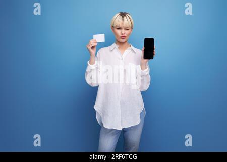giovane segretaria bionda caucasica vestita con una camicia bianca contiene una tessera di plastica e uno smartphone con un mockup. concetto aziendale Foto Stock