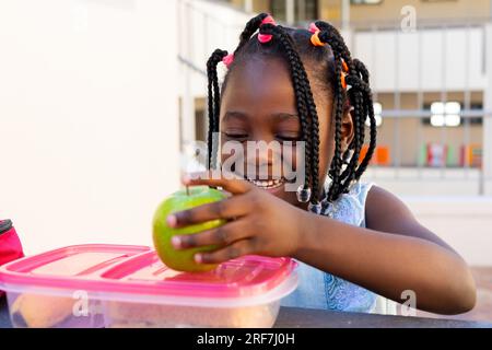 Studentessa afroamericana a tavola e pranzando sano con mela fuori dalla scuola elementare Foto Stock