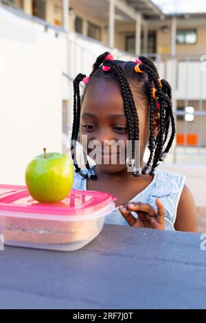 Studentessa afroamericana a tavola e pranzando sano con mela fuori dalla scuola elementare Foto Stock