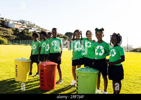 Ritratto di bambini felici e diversificati che raccolgono ricicli abbracciano nel soleggiato campo della scuola elementare Foto Stock