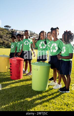 Ritratto di bambini felici e diversificati che raccolgono ricicli abbracciano nel soleggiato campo della scuola elementare Foto Stock