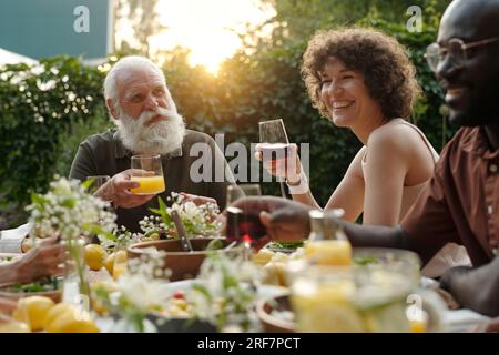 Giovane donna felice con un bicchiere di succo d'arancia seduta tra due uomini al tavolo servito e tifa mentre chiacchiera con altri membri della famiglia Foto Stock