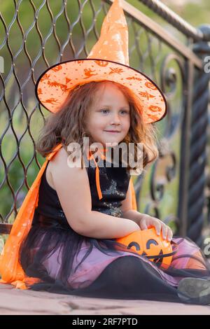 Una graziosa bambina in costume da strega per Halloween cammina nel parco in un cesto di caramelle a forma di zucca. Foto di alta qualità Foto Stock