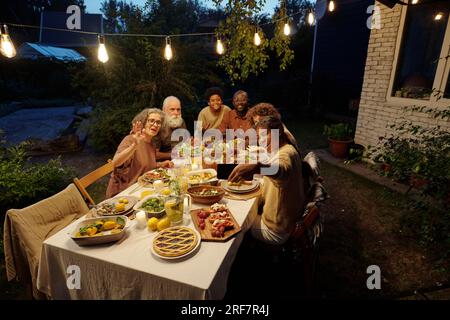 Ragazza felice che scatta selfie con una grande famiglia interculturale sul suo smartphone mentre si siede tra i genitori e i nonni a bordo di un tavolo servito Foto Stock