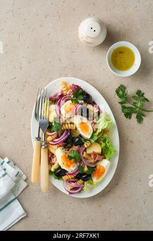 Insalata di tonno con pasta, uova, patate, olive, cipolle rosse e salsa in piatto bianco su vecchio fondo di cemento grigio chiaro. Insalata Nicoise. Francese Foto Stock