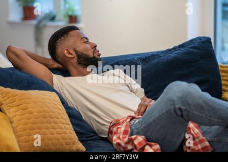 Il ragazzo afro-americano sognante e premuroso siede su divani appoggiati nel monolocale. Foto Stock