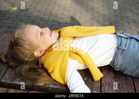 Una bambina bionda in età prescolare in jeans, un dolcevita e un maglione si trova su una panchina con gli occhi chiusi e gode del tempo. Pho orizzontale Foto Stock