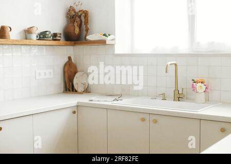 Cucina dal design moderno e minimalista. Eleganti armadi da cucina bianchi con rubinetto in ottone, ripiani in granito e ripiani in legno con utensili in un nuovo scandinav Foto Stock