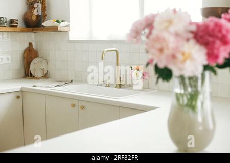Cucina dal design moderno. Elegante rubinetto in ottone e lavandino in granito bianco, armadietti da cucina con pomelli e bouquet di peonie nella nuova casa scandinava. Moderno Foto Stock