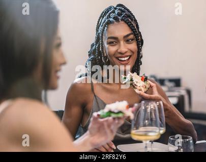 Donna afro-americana che tiene una fetta di pizza e chiacchiera con i suoi amici. Una figura sfocata di una donna in primo piano. Tutti stanno sorridendo in questo Foto Stock