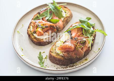 Toast a colazione con formaggio cremoso, salmone, olio e rucola su un piatto, sfondo bianco, primo piano. Ricetta per la dieta Keto. Foto Stock