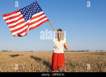 La donna sta in piedi con la schiena e porta con orgoglio la grande bandiera americana nel campo di grano illuminato dalla luce del sole. Giorno dell'indipendenza degli Stati Uniti d'Amer Foto Stock