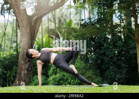 Donna asiatica pratica yoga facendo Utthita Trikonasana esercizio, esteso triangolo posa sul tappeto accanto a un lago nel parco all'aperto. Foto Stock