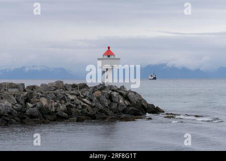 Piccolo faro, alla fine di un muro di banchina, e traghetto per Gryllefjord visto da una barca. Andenes, Andøya, Arcipelago di Vesterålen, Nordland, Norvegia Foto Stock