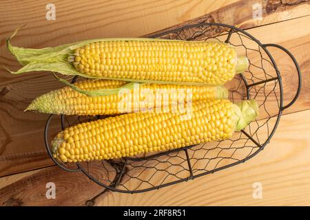 Tre pannocchie di mais dolce biologico in un cesto di metallo, primo piano, su un tavolo di legno, vista dall'alto. Foto Stock