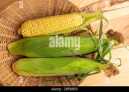 Tre pannocchie di mais dolce biologico in un piatto di paglia, primo piano, su un tavolo di legno, vista dall'alto. Foto Stock