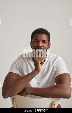 Ritratto minimalista di un uomo nero sorridente che posa in studio su sfondo bianco seduto all'indietro sulla sedia Foto Stock